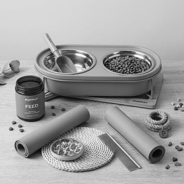 Purrpurr pet feeding and mealtime accessories in a grayscale flat lay, featuring a double elevated feeding station with stainless steel bowls, a food storage jar, silicone feeding mats, a slow feeder bowl, and a wooden food scoop on a light tabletop.