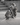 A grayscale outdoor shot of a small dog and a cat sitting together on stone steps. Both pets are wearing matching insulated, hooded winter parkas with utility harnesses and a "PurrPurr" brand patch on the side.